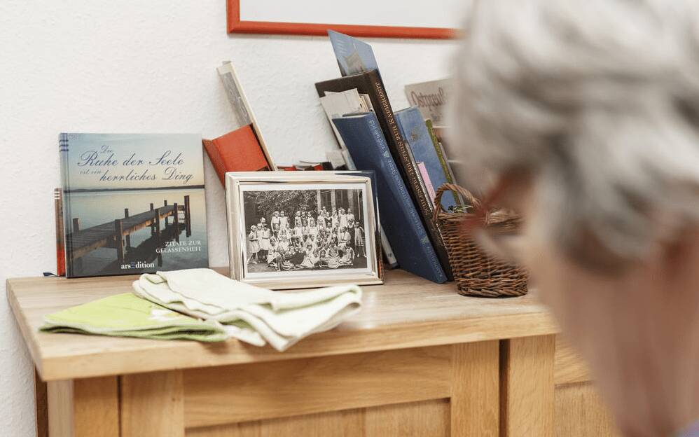 Bewohnerin im Profil vor einer Kommode mit Erinnerunsgstücken und Bildern in ihrem Zimmer