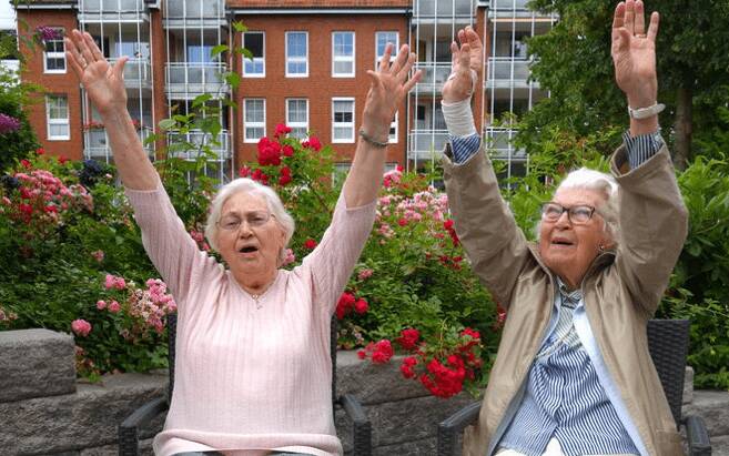Zwei alte Damen sitzen auf der Terrasse, lachen und machen Faxen.