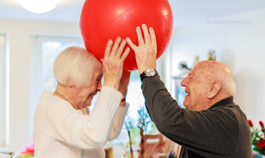 Senior und Seniorin halten Ball über den Köpfen gemeinsam fest und strahlen sich an