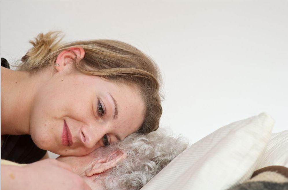 Junge Frau legt ihre Wange an die einer bettlägerigen Seniorin