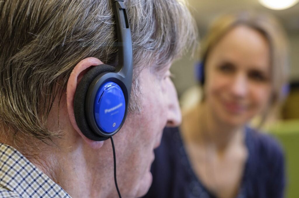 Senior hört Musik über Kopfhörer. Eine junge Dame schaut zu.